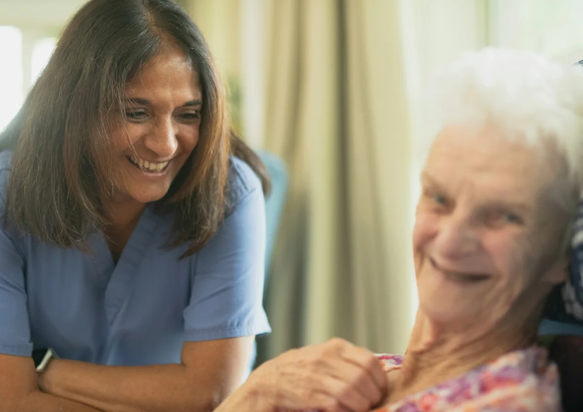 Smiling carer in a blue tunic sharing a laugh with an older woman