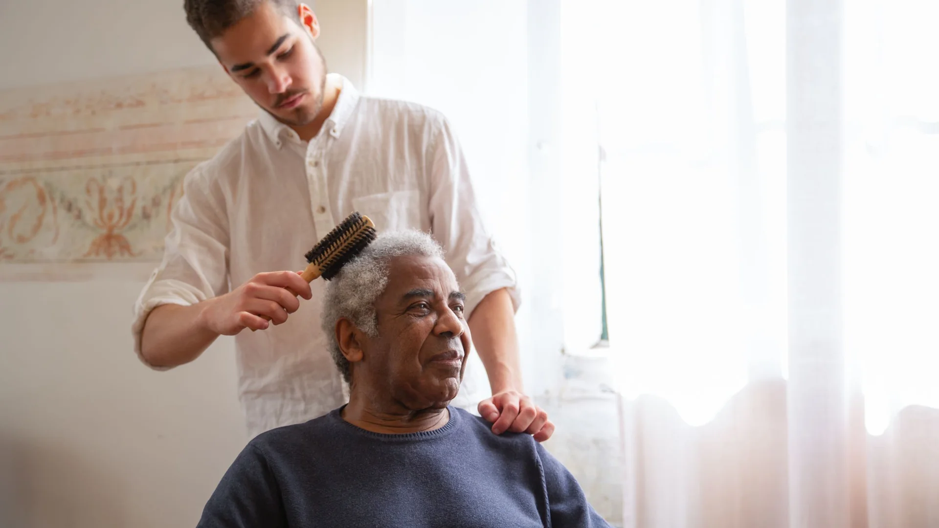 Young male carer brushing an older man's hair in a bright living room