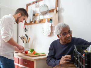 Carer preparing fresh vegetables in the kitchen while an older man looks on