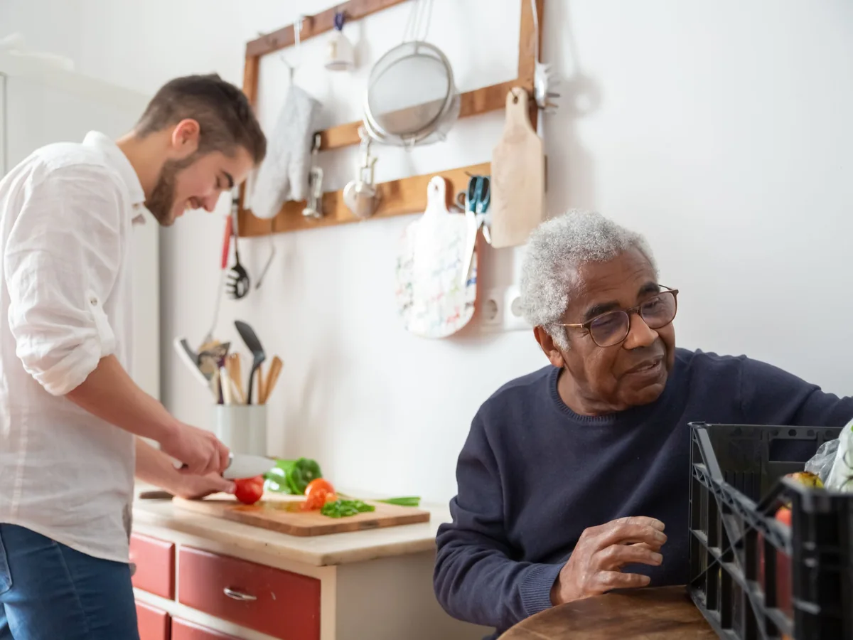 Carer preparing fresh vegetables in the kitchen while an older man looks on