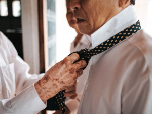 Hands helping an older man fasten his tie while he gets dressed