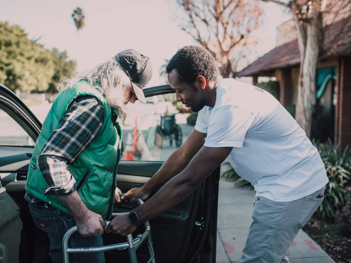 Helper steadying a walking frame as an older man gets out of a car